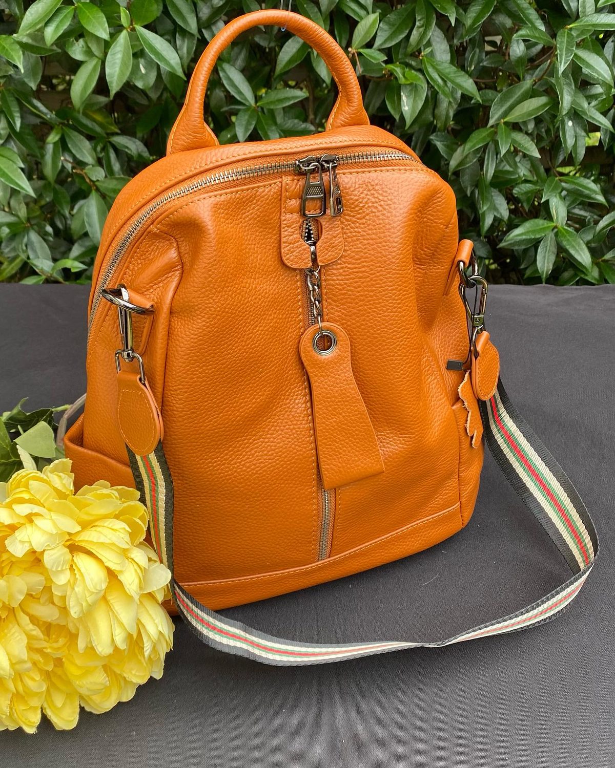 Tan pebbled leather backpack with a striped strap, photographed against greenery